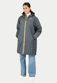 Manteau imperméable gris avec capuche, fermeture éclair jaune à l'avant et poignets élastiques. Porté par-dessus un jean large bleu et des chaussures blanches.