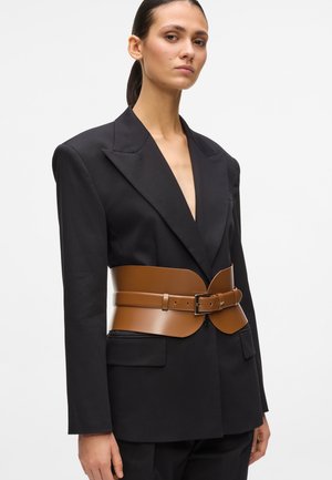 Femeie purtând un blazer negru supradimensionat, cu o centură corset largă din piele maro care strânge talia, stând în fața unui fundal alb simplu.