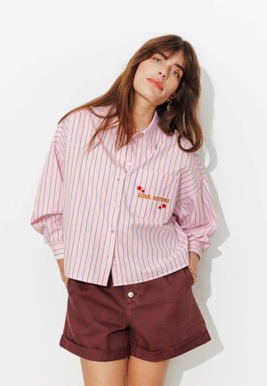 Femme portant une chemise rose à rayures oversize avec le texte "SOUL SISTERS" sur la poche et un short bordeaux roulé, debout devant un fond blanc.