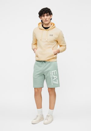 Sudadera amarilla con capucha y bolsillo canguro, junto con pantalones cortos verde menta con texto gráfico blanco. Zapatillas claras y calcetines blancos.