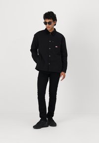 Cămașă neagră cu guler, buzunar frontal și detaliu cu logo. Asortată cu blugi negri și adidași asortați. Ochelarii completează look-ul.