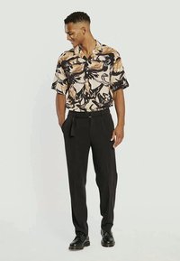 Camisa de mangas curtas em bege com padrões abstratos em preto e castanho, combinada com calças pretas e sapatos pretos. Tecido suave, corte relaxado.
