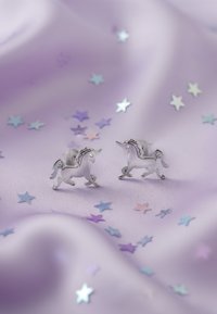 Boucles d'oreilles en argent en forme de licorne sur un tissu lavande, entourées de petites étoiles pastel. Les boucles d'oreilles présentent des détails complexes sur les crinières.