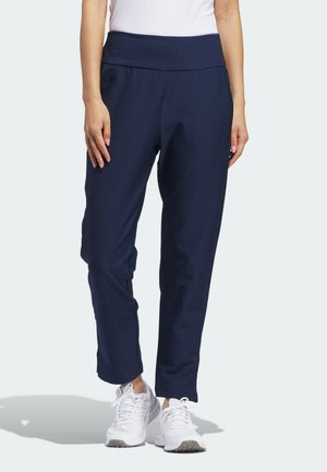 Vrouw draagt marineblauwe broek en witte sneakers, staand tegen een effen lichte achtergrond.