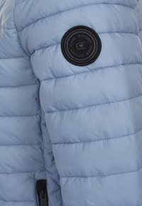 Világoskék puffernadrág, steppelt textúrával, fekete, kör alakú logópánttal az ujjon és cipzáras részlettel az alján.