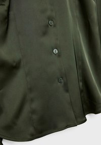 Camicia di raso verde scuro con una consistenza liscia, caratterizzata da sei bottoni circolari sulla parte anteriore e un orlo curvato.