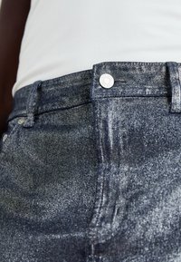 Džínsy z denimu s tmavomodrou, mierne metalickou povrchovou úpravou. Obsahujú klasický dizajn s piatimi vreckami a kovovým zapínaním.