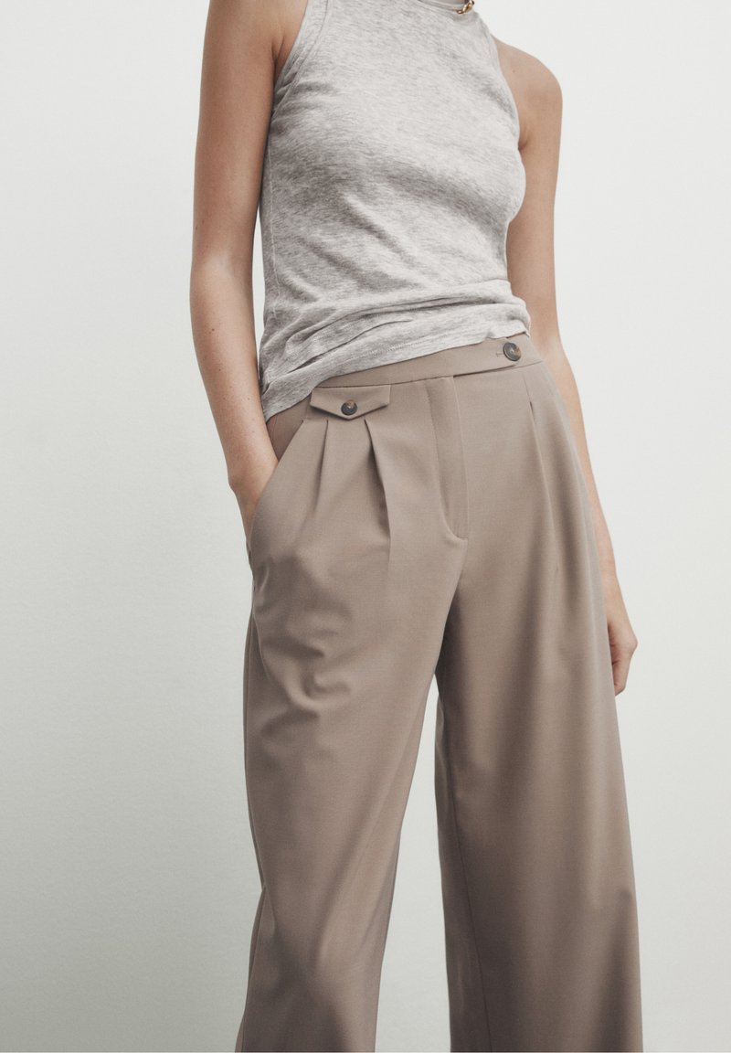 Haut sans manches gris clair associé à un pantalon beige taille haute plissé, avec deux poches avant et des boutonnières en décoration.
