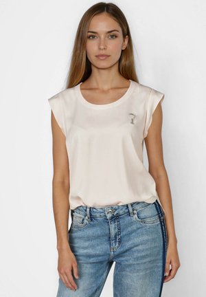 Young woman wearing sleeveless pale pink blouse with small silver brooch and blue denim jeans, standing against plain background.