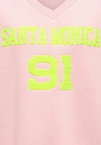 Tissu rose à col en V avec des lettres texturées vert-jaune indiquant « SANTA MONICA » et le numéro « 91 » en dessous.