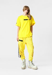 Camisa amarilla de gran tamaño con una etiqueta negra, combinada con pantalones jogger amarillos que tienen una impresión lateral, y botas blancas con cordones.