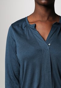 Türkisfarbende Bluse mit langen Ärmeln und V-Ausschnitt, ausgestattet mit kleinen Falten am Kragen und einem einzelnen Knopfverschluss. Glatte Stofftextur.