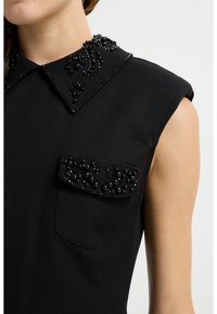 Schwarze ärmellose Bluse mit verziertem Kragen, der mit schwarzen Perlen und Pailletten geschmückt ist, sowie einer dekorativen Tasche mit ähnlichen Perlendetails.