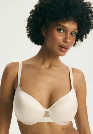 Kobieta o kręconych włosach, nosząca gładki, jasnobeżowy biustonosz z cienkimi ramiączkami, ukazująca naturalny odcień skóry i subtelne tatuaże na obu ramionach.