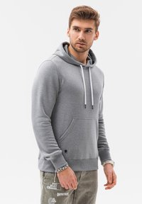 Felpa grigia chiara realizzata in materiale morbido, con tasca frontale, cordini e polsini a costine. Il design è semplice e funzionale.