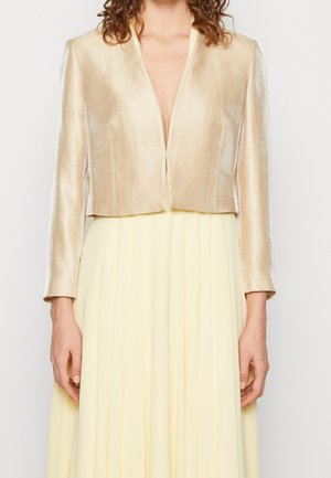 Vrouw die een glanzend beige cropped blazer met lange mouwen draagt over een lichtgele geplooide jurk, getoond van borst tot heupen.