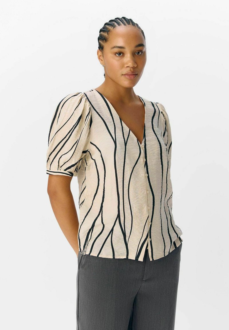 Blouse beige en V avec des rayures noires ondulées, manches bouffantes et boutons sur le devant. Le tissu a une finition texturée.