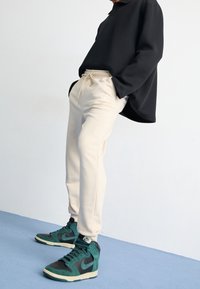 Krémsvětlé tepláky se stahovací šňůrkou a postranními kapsami, v páru s černými a petrolejově modrými kotníkovými teniskami. Černé tričko s dlouhým rukávem. Minimalistické prostředí.