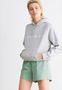 Sivi dukserica s kapuljačom i prednjim džepom, sa izvezenim logotipom "carhartt", u kombinaciji sa svijetlozelenim šortsevima koji imaju bijeli logotip.