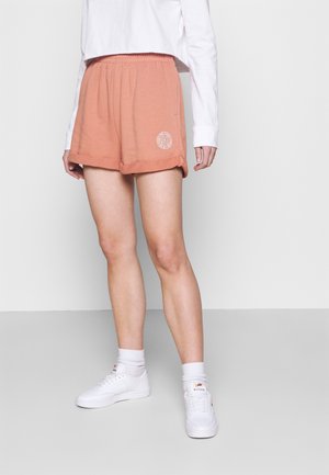 Shorts en coton de couleur pêche avec une taille élastique, présentant un logo blanc en bas et des ourlets roulés, associés à des baskets blanches.