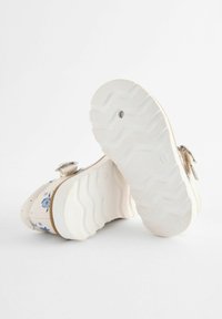 Beige Schuhe mit floralem Muster, dicken weißen Sohlen und einer Schnallenverzierung. Die Sohle hat einen strukturierten Grip mit einem Wellenmuster.