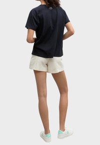 Černé bavlněné tričko s krátkým rukávem a uvolněným střihem, s detailem švu na zadní straně, spárované se světle béžovými šortkami a bílými teniskami s mátovými akcenty.