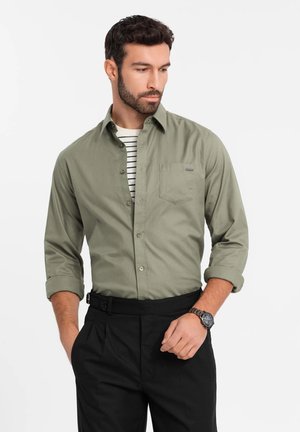 Camisa verde oliva de botón, con un bolsillo en el pecho, combinada con un cinturón negro y pantalones oscuros de vestir. Presenta mangas enrolladas y una camiseta interior a rayas.
