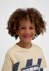 Lys beige t-shirt med et stort blåt grafisk design. Barn med krøllet, brunt hår og et stort smil, der viser et hul i deres tænder.