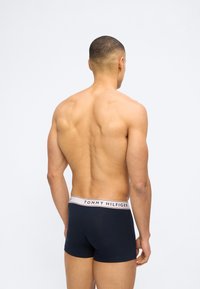 Μυώδης άνδρας με κοντά μαλλιά, γυρισμένος πλάτη, φορώντας σκούρο μπλε μποξεράκια Tommy Hilfiger με λευκή ζώνη μέσης και κείμενο.