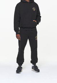 Completo felpa nera con hoodie e pantaloni sportivi, dotato di tasca frontale, logo ricamato in oro, polsini e orli delle caviglie a coste. Abbinato a sneakers nere.