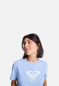 Světle modré bavlněné tričko s bílým a neonově zeleným logem srdce a názvem značky "ROXY" pod ním. Krátké rukávy a klasický střih.