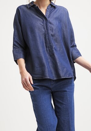 Kvinde iført løs mørkeblå knapbluse med trekvartærmer og matchende blå jeans mod en ensfarvet lys baggrund.