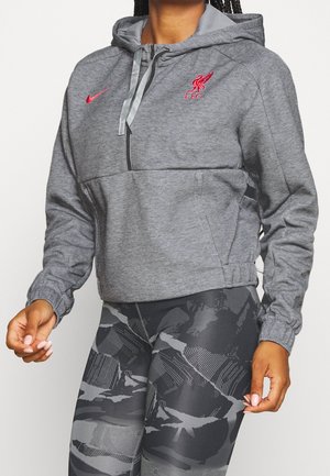 Person trägt einen grauen Liverpool FC Hoodie mit roten Logos und schwarz-grau gemusterten Leggings vor weißem Hintergrund.