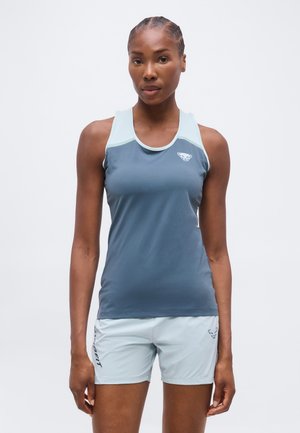 Femme portant un débardeur de sport bleu sans manches et un short de sport bleu clair, debout devant un fond uni.