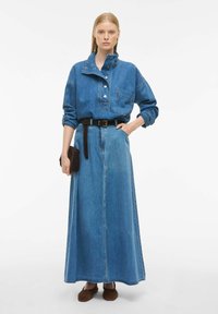 Ženska, oblečena v modro platneno srajco z gumbi in zavihanimi rokavi, ujemajočo dolgo denim krilo, rjav pas, rjave čevlje in drži majhno rjavo torbico.