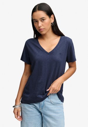 Femme aux longs cheveux foncés portant des boucles d'oreilles créoles, un t-shirt bleu marine à col en V, un jean bleu clair et des bracelets argentés.