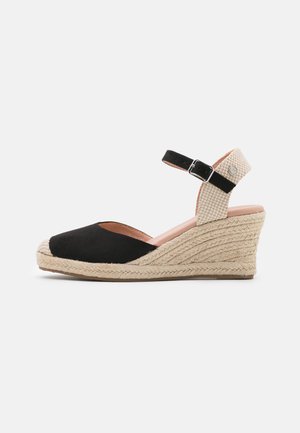 Sandalia de cuña negra y beige con suela de esparto tejida, correa en el tobillo con hebilla plateada y diseño de punta cerrada.