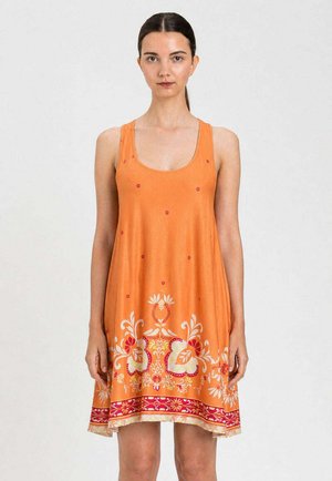 Donna che indossa un vestito arancione senza maniche con motivi floreali e bordo decorativo, in piedi davanti a uno sfondo semplice.