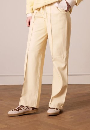 Yellow wide-leg trousers with a smooth texture and side seams. Paired with brown and cream sneakers featuring a spotted design.