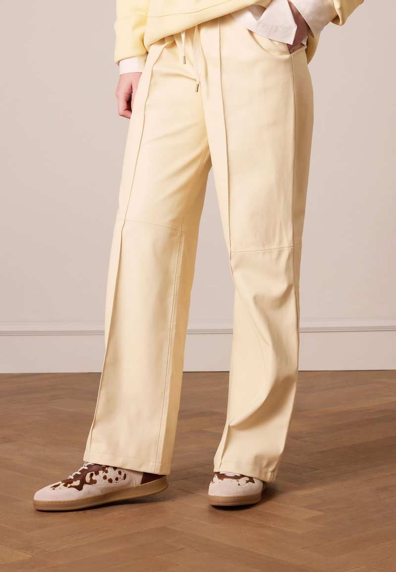 Yellow wide-leg trousers with a smooth texture and side seams. Paired with brown and cream sneakers featuring a spotted design.