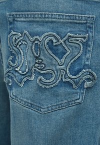 Blugi din denim albastru, cu un design decorativ brodat în albastru deschis pe buzunarul din spate, cu margini desfăcute și variații de textură.