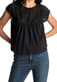 Femme portant un blouse en dentelle noire à manches courtes associée à un jean en denim bleu, les mains posées sur les hanches.