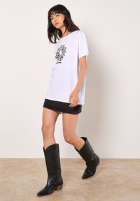 Weißes Baumwoll-T-Shirt mit einem schwarzen geometrischen Druck, kombiniert mit einem schwarzen Denim-Minirock und kniehohen schwarzen Leder-Stiefeln im Cowboy-Stil.