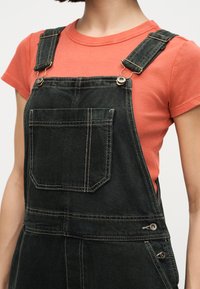 Γυναίκα που φορά ξεβαμμένη μαύρη τζιν φόρμα με μπροστινή τσέπη πάνω από κοραλλί κοντομάνικη ριμπ μπλούζα, εμφανίζοντας τον πάνω κορμό και τα λουριά.