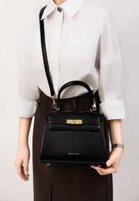 Czarna skórzana torebka o wyraźnym kształcie, z złotymi elementami, górną rączką i odpinanym paskiem na ramię. Posiada przedni klapek oraz logo.