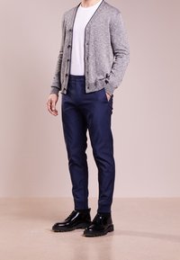 Man med grå knapp-up cardigan, vit t-shirt, marinblå slim-fit byxor och svarta polerade läder snörskor, stående med en hand i fickan.