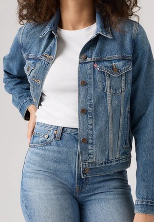 Woman wearing light blue denim jacket and jeans with white t-shirt, hand resting on hip, plain background.