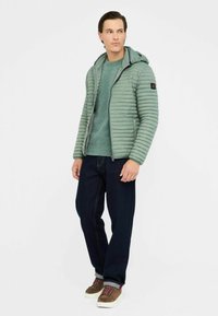 Giacca puffer verde chiaro con cuciture orizzontali e cappuccio, indossata sopra un maglione verde a trama, abbinata a jeans scuri e sneakers marroni.