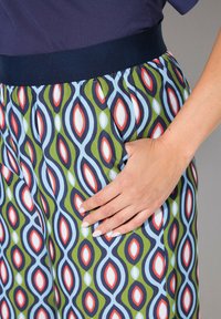 Hand rustend in de zak van een broek met een marineblauwe tailleband en een geometrisch patroon in groen, blauw, rood en wit.