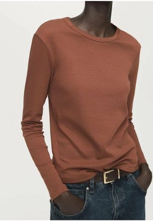 Person iført rustfarvet langærmet skjorte stoppet ned i blå jeans med sort bælte, hænder i lommerne, mod en ensartet lys baggrund.